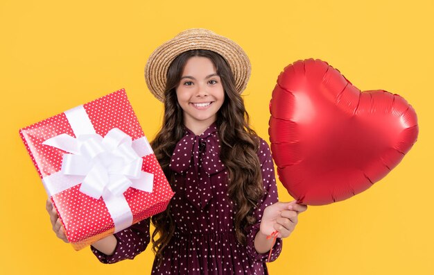 Menina adolescente positiva com balão de coração vermelho e caixa de presente em fundo amarelo