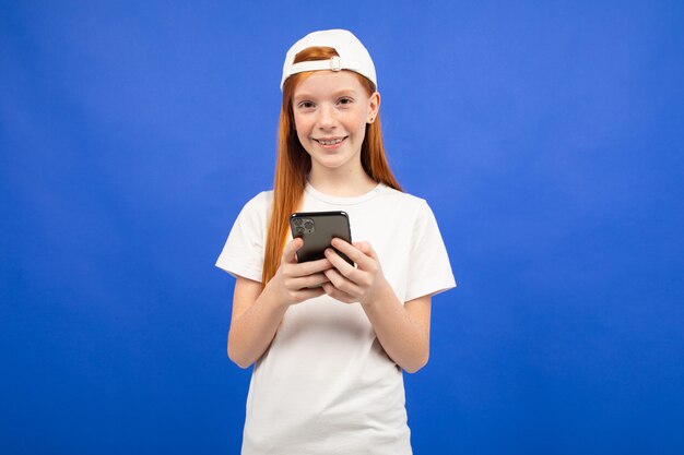 Menina adolescente ruiva alegre em uma camiseta branca se comunica nas redes sociais a partir de um smartphone em um azul