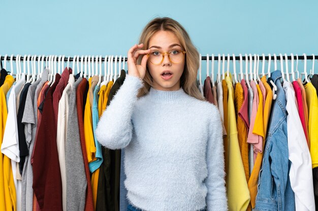 Menina adolescente russa comprando roupas isoladas em azul com óculos e surpresa