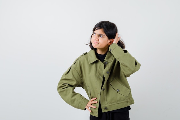Menina adolescente segurando a mão atrás da orelha, olhando para longe na jaqueta verde do exército e parecendo curiosa. vista frontal.