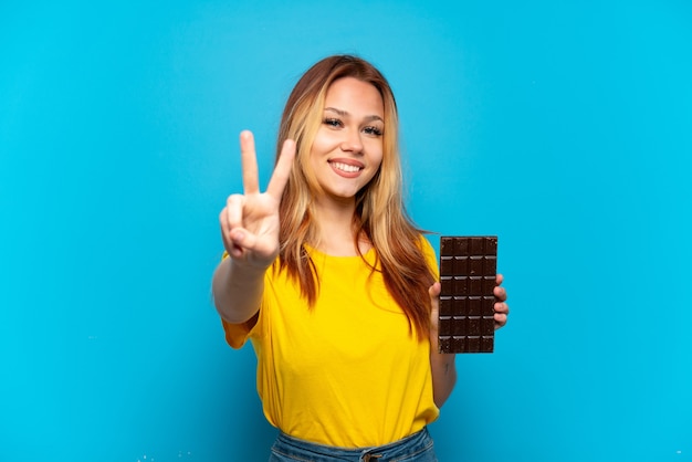 Menina adolescente segurando chocolate sobre fundo azul isolado, sorrindo e mostrando o sinal da vitória