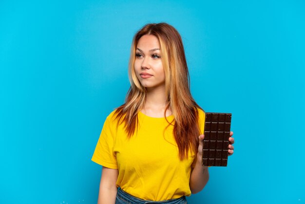 Menina adolescente segurando chocolate sobre um fundo azul isolado, olhando para o lado
