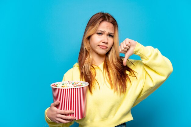 Menina adolescente segurando pipocas sobre um fundo azul isolado, mostrando o polegar para baixo com expressão negativa