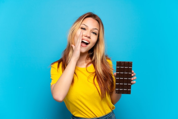 Menina adolescente segurando um chocolate sobre um fundo azul isolado e gritando com a boca bem aberta