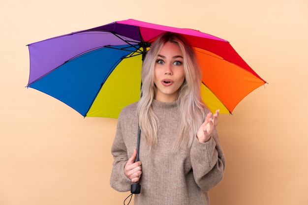 Menina adolescente segurando um guarda-chuva sobre parede isolada com expressão facial de surpresa