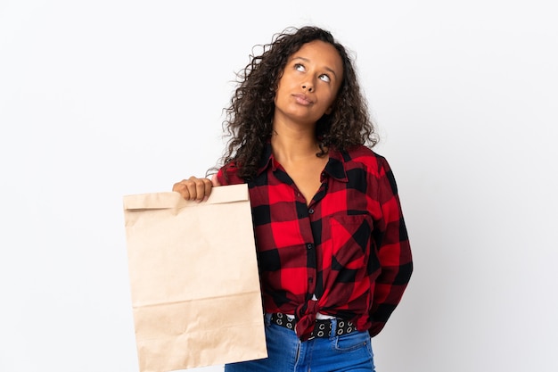 Menina adolescente segurando uma sacola de compras de supermercado para levar isolada na parede branca e olhando para cima