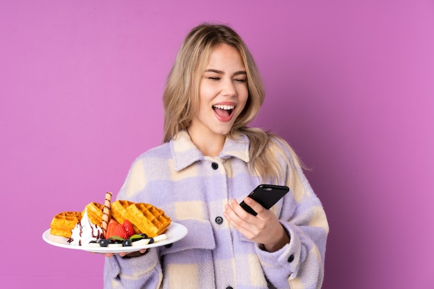 Menina adolescente segurando waffles isolados em roxo com o telefone em posição de vitória