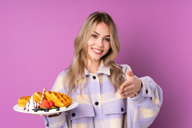 Menina adolescente segurando waffles na parede roxa, apertando as mãos para fechar um bom negócio