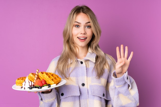 Menina adolescente segurando waffles na parede roxa feliz e contando quatro com os dedos