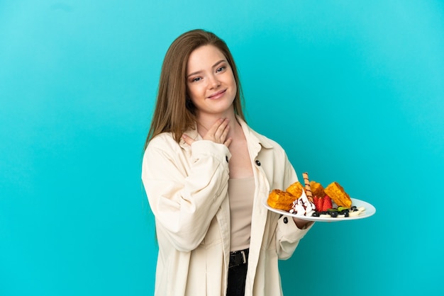 Menina adolescente segurando waffles sobre um fundo azul isolado, olhando para cima enquanto sorri