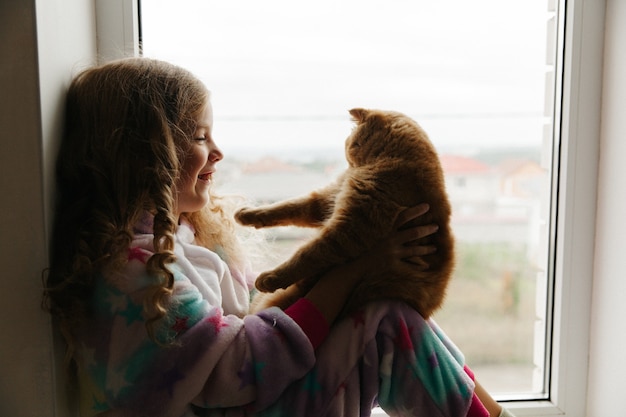 Menina adolescente senta-se de pijama na janela da casa e segura um gato ruivo. fique em casa.