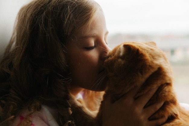 Foto menina adolescente senta-se de pijama na janela da casa e segura um gato ruivo. fique em casa.