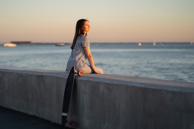 Menina adolescente skatista no pôr do sol à beira-mar. Jovem aproveita a noite de verão no rio ou no mar, olhando para a água