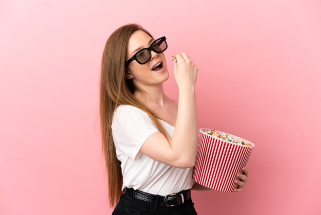 Menina adolescente sobre fundo rosa isolado com óculos 3D e segurando um grande balde de pipocas