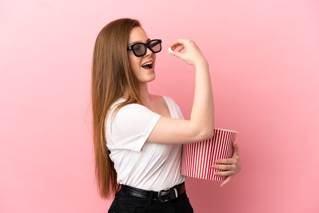 Menina adolescente sobre fundo rosa isolado com óculos 3D e segurando um grande balde de pipocas