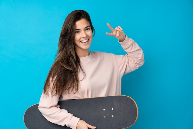 Menina adolescente sobre parede azul isolada com um skate com expressão feliz