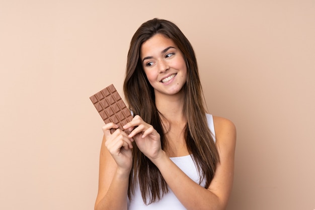 Menina adolescente sobre parede isolada, tomando um comprimido de chocolate e feliz