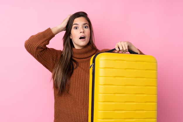 Menina adolescente sobre parede rosa isolada em férias com mala de viagem e surpreso
