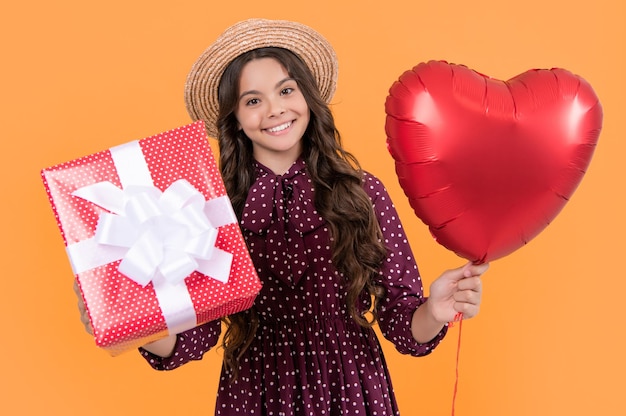 Menina adolescente sorridente com balão de coração vermelho e caixa de presente em fundo amarelo