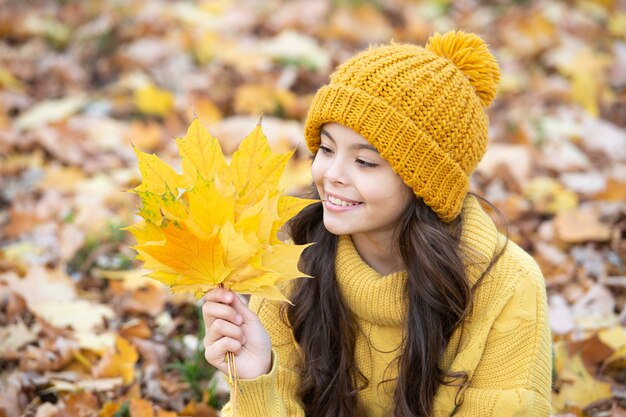 Menina adolescente sorridente com chapéu amarelo segurar folha de plátano ao ar livre no parque, outono.