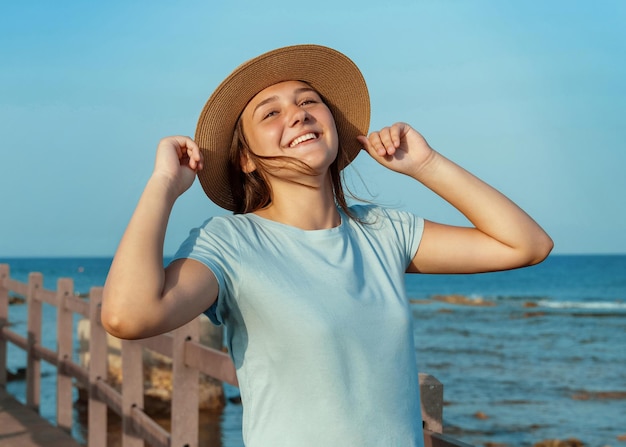 Menina adolescente sorridente em pé no cais de madeira à beira-mar ao pôr do sol, vestindo uma camiseta azul claro e tocando com as duas mãos seu chapéu de palha. viagem de verão, perto do conceito de natureza. maquete de camiseta