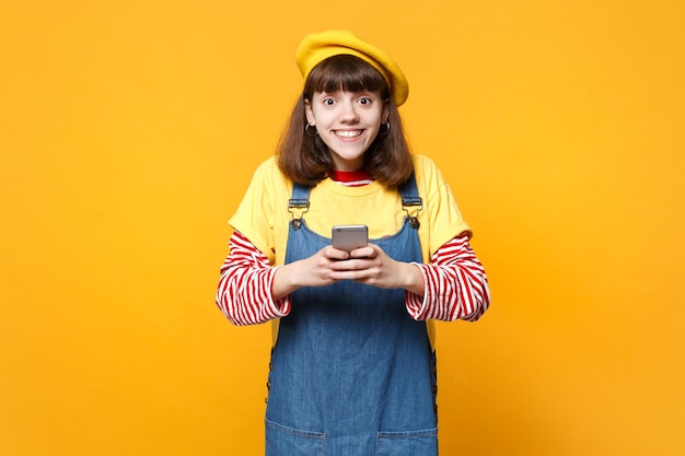 Menina adolescente sorridente na boina francesa, vestido jeans usando telefone celular, digitando mensagem sms isolada em fundo amarelo no estúdio. Emoções sinceras de pessoas, conceito de estilo de vida. Mock up espaço de cópia.