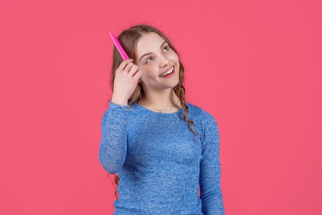 Menina adolescente sorridente, penteando o cabelo encaracolado com escova de cabelo no fundo rosa