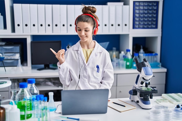 Menina adolescente trabalhando no laboratório cientista sorrindo feliz apontando com a mão e o dedo para o lado
