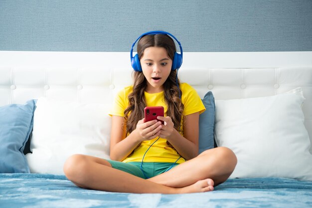 Menina adolescente usando fones de ouvido assistindo vídeos no smartphone sentado na cama em seu quarto Rosto surpreso surpreende emoções de adolescente