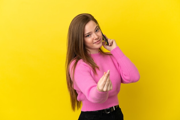 Menina adolescente usando telefone celular sobre fundo amarelo isolado fazendo gesto de dinheiro