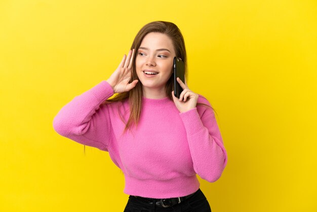 Menina adolescente usando telefone celular sobre fundo amarelo isolado, ouvindo algo colocando a mão na orelha