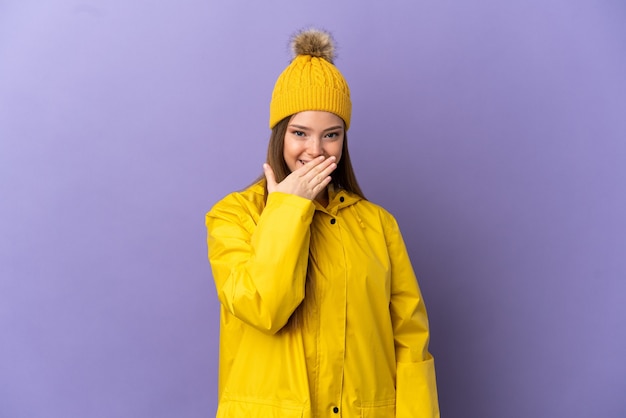 Menina adolescente vestindo um casaco à prova de chuva sobre um fundo roxo isolado, feliz e sorridente, cobrindo a boca com a mão