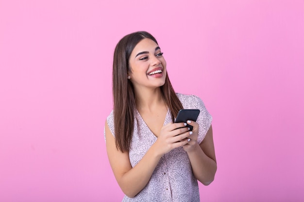 Menina adorável com telefone móvel na mão expressando emoções positivas Mulher atraente com camisa rosa segurando smartphone e enviando mensagens de texto em fundo rosa de estúdio