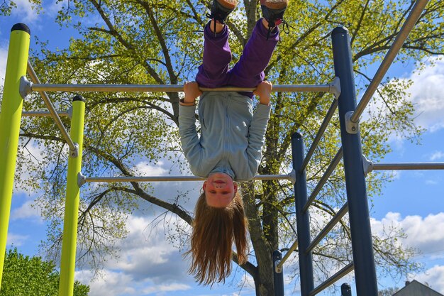 Foto menina adorável rindo felizmente pendurada de cabeça para baixo em barras paralelas no playground