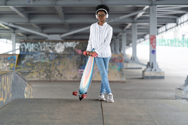 Menina africana com skate feminino na moda vestida com roupa urbana e fones de ouvido segurando longboard