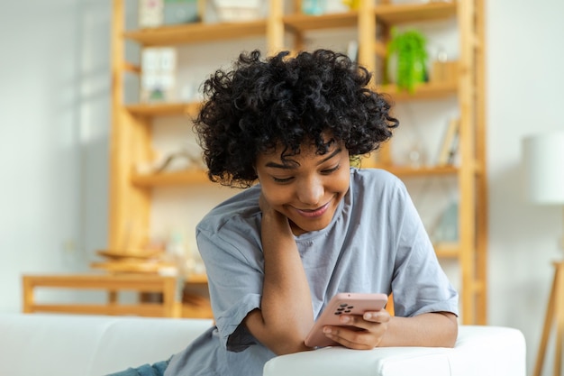 Menina africana segurando tela sensível ao toque do smartphone digitando página de rolagem em casa Mulher com celular navegando na internet usando aplicativos de mídia social jogando jogo Compras online Notícias da Internet vício em celular