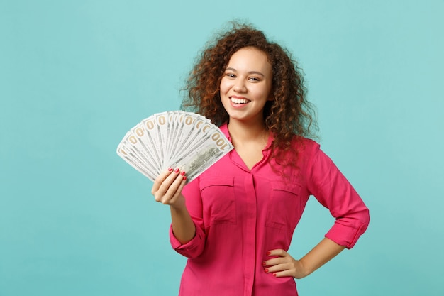 Menina africana sorridente em roupas casuais, segurando um leque de dinheiro nas notas de dólar, dinheiro isolado no fundo da parede azul turquesa. emoções sinceras de pessoas, conceito de estilo de vida. simule o espaço da cópia.