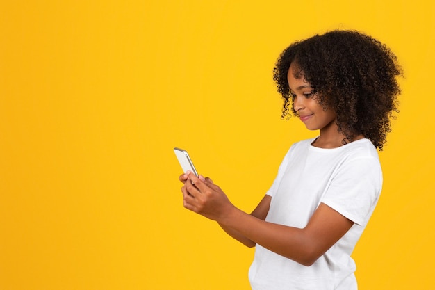 Menina afro-americana adolescente encaracolada sorridente em camiseta branca digitando no telefone