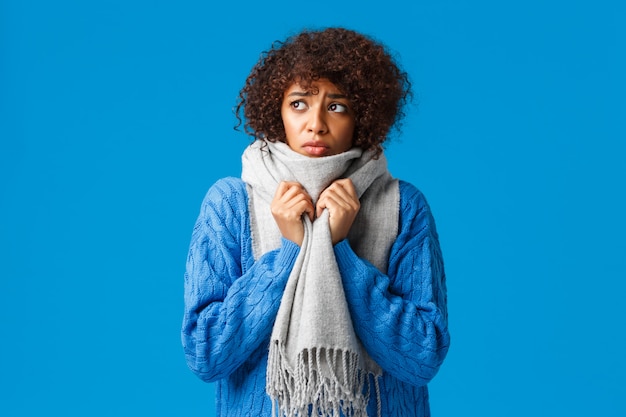 Menina afro-americana boba e triste, sombria, olhando para fora do inverno com vento e vento, enrole o pescoço com um lenço quente, tremer de frio sentindo desconforto por causa da baixa temperatura em casa, azul