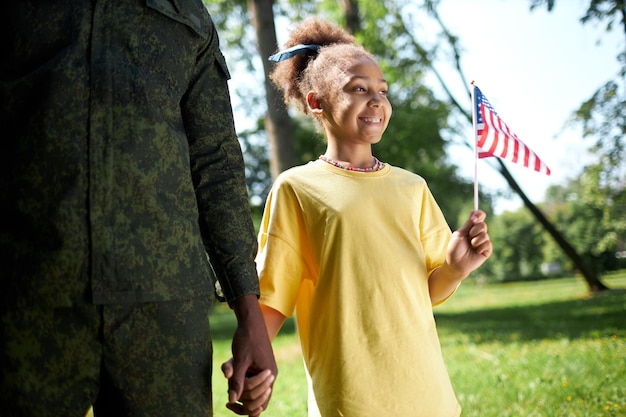 Menina afro-americana com bandeira americana andando com o pai em uniforme militar ao ar livre