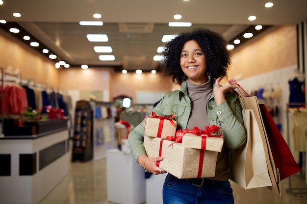 Menina afro-americana comprando presentes no shopping na venda de natal Conceito de férias de ano novo Mulher racial mista atraente sorridente com caixas de presente de papel colorido usando chapéu de natal na loja ou loja