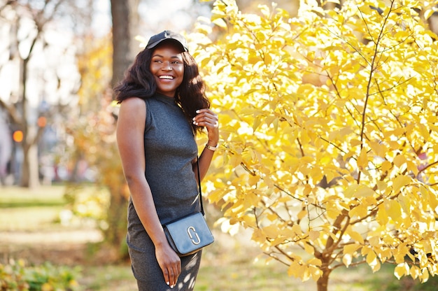 Menina afro-americana elegante na túnica cinza, saco crossbody e boné posou em dia ensolarado de outono contra folhas amarelas. mulher modelo áfrica.