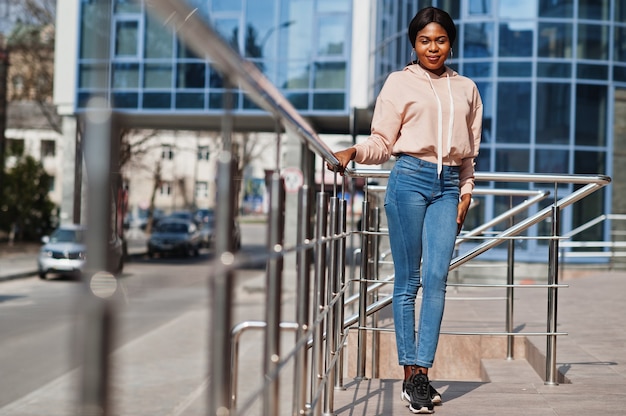 Menina afro-americana hippie com capuz rosa, jeans posando na rua contra o prédio com janelas azuis.