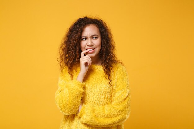 Menina afro-americana preocupada em suéter de pele posando isolado no fundo da parede laranja amarela, retrato de estúdio. conceito de estilo de vida de pessoas. mock up espaço de cópia. olhando para o lado, roendo as unhas.