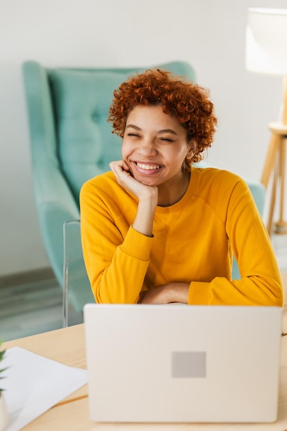 Foto menina afro-americana sorridente usando laptop no escritório em casa, digitando, conversando, lendo, escrevendo e-mail.