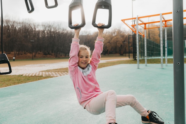 Menina alegre brinca no playground. A criança está envolvida em argolas de ginástica na rua. A criança pratica esportes em simuladores.