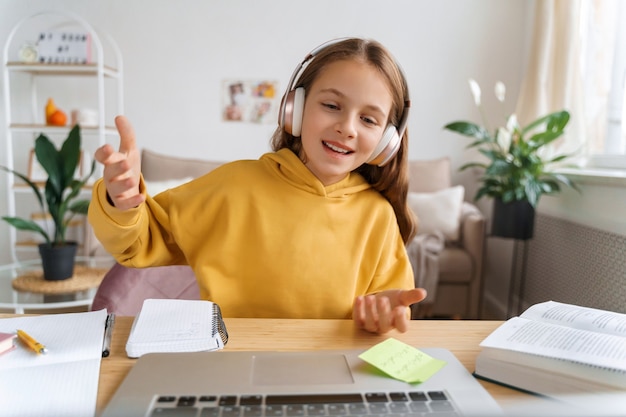 Foto menina alegre com fones de ouvido, sentada no quarto dela, falando para a câmera, fazendo uma videochamada, usando um laptop para se comunicar com um amigo