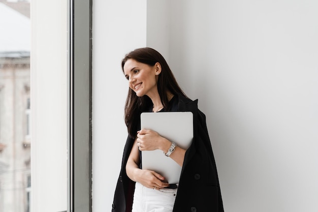 Menina alegre com laptop parado perto de uma janela grande no escritório branco Jovem mulher está abraçando seu laptop e sorrindo no café
