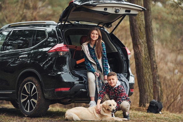 Menina alegre com seu irmão senta-se na parte de trás do carro moderno com seu cachorro ao ar livre na floresta