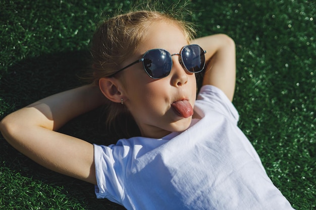 Menina alegre de 8 anos em óculos de sol encontra-se na grama no parque Criança ao ar livre descansa na vista do parque de verão de cima Lugar para uma inscrição